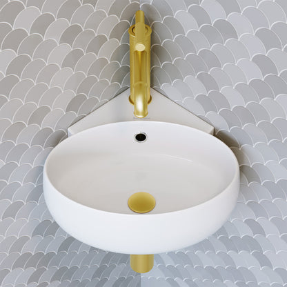 Round wall-hung bathroom sink in white with gold drain cover and gold faucet against gray scale tile wall.