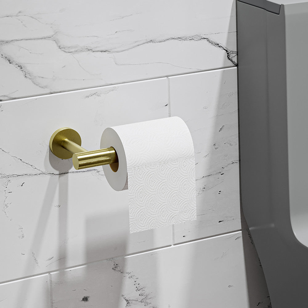 Avallon toilet paper holder in brushed gold mounted on white tiled wall next to dark gray toilet.